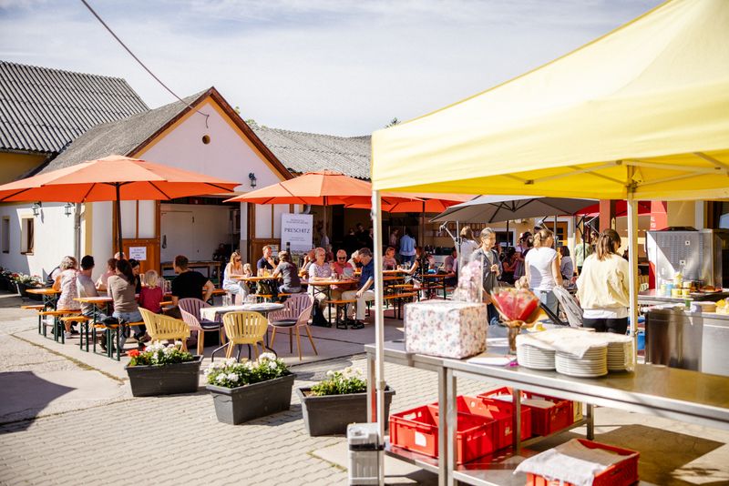 Hof von Irene’s Bauernladen bei der Jubiläumsfeier: Gäste unter orangen Sonnenschirmen, daneben ein gelbes Verkaufszelt