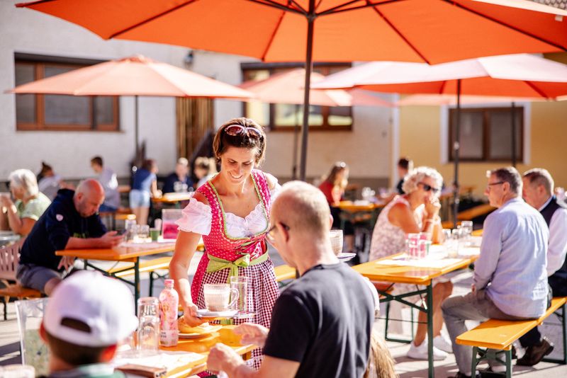 Gäste an Biertischen unter orangen Sonnenschirmen im Hof, eine Mitarbeiterin im Dirndl serviert
