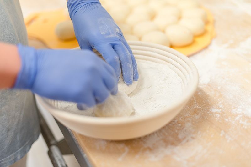 Hände in blauen Handschuhen bemehlen Teig in einem Gärkorb
